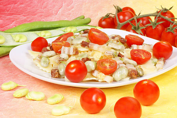 Farfalle with broad beans, bacon and fresh cherry tomatoes on a white plate. Studio shot over red-yellow background
