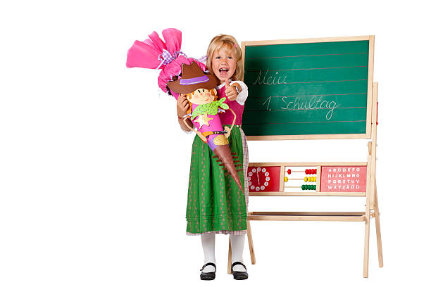 Girl on first school day stands beside chalk board and shows thumb. Isolated on white background.