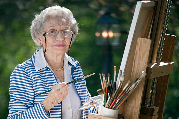 A smiling senior woman is in an outdoor setting painting. Horizontal shot.