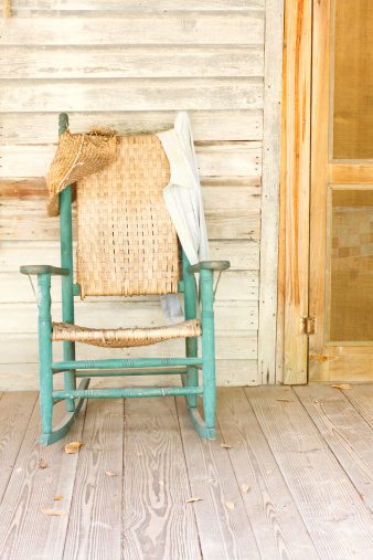 A vintage green rocking chair in a rural setting on the front porch of an old farmhouse.