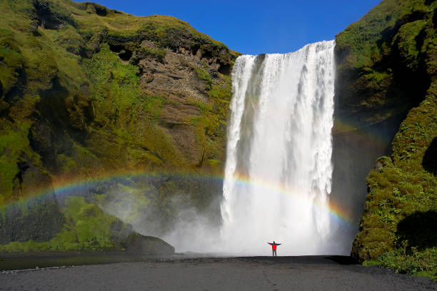 Skogafoss waterfall Man in a red jacket stood under a rainbow appears across the base of the huge Skogafoss waterfall in South Iceland skogafoss stock pictures, royalty-free photos & images