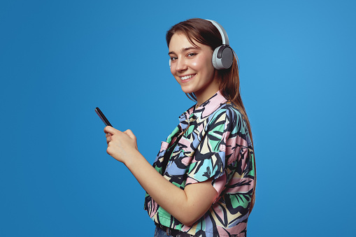 Side view photo of charming woman in headphones listening to music and using mobile phone, isolated over blue background. People, lifestyle and emotion concept