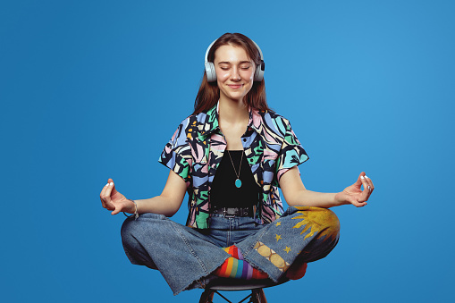 Pretty smiling woman in stylish clothes sitting on chair in lotus pose, meditating and listening a peaceful song on headphones, isolated on blue background. Relaxation, leisure activities concept