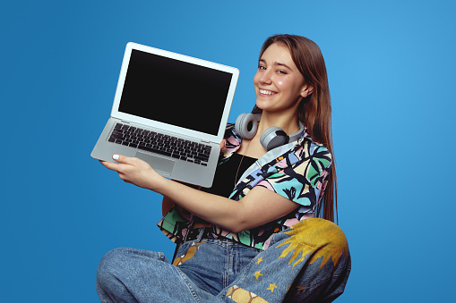 Young student girl holding laptop while sitting with legs crossed on chair, showing empty black screen over blue background. Computing devices concept