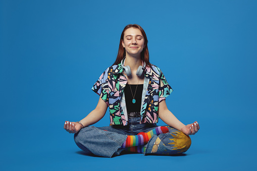 Calm young girl with headphones wearing stylish colorful clothes, meditating with eyes closed while practicing mindfulness, sitting on floor in lotus pose, isolated over blue background