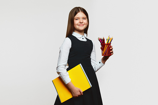 Little pupil girl in school uniform holding notebook and colorful pencils, looking at camera while standing against white background