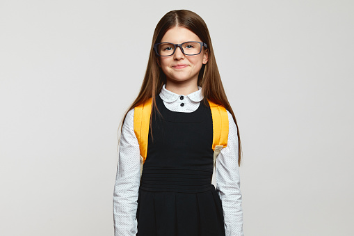 Good looking nerdy pupil kid wearing eyeglasses, school uniform and yellow backpack staring at camera against white background. Back to school concept