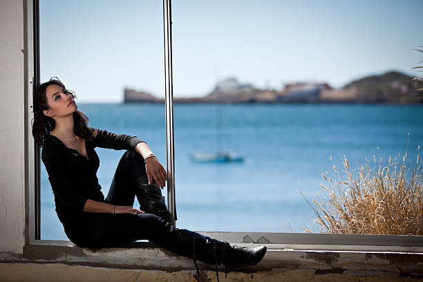 Pretty young brunette woman sitting in a window overlooking the ocean with a sailboat blurred in the background