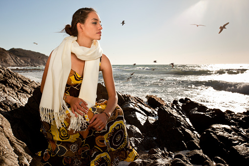 Pretty young brunette woman sitting on a rocky beach at sunset with seagulls flying overhead