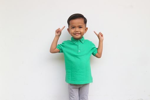 A smiling toddler boy looks at the camera raising his hands and pointing up, isolated on white background