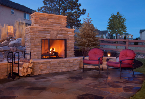 A backyard patio features a fireplace and casual furnishings.  The outdoor fireplace burns with a bright orange glow and is covered by a metal grate.  There is extra wood stored to the left of the stone fireplace in a black unit.  There are two metal chairs with red cushions placed on the flagstone patio.