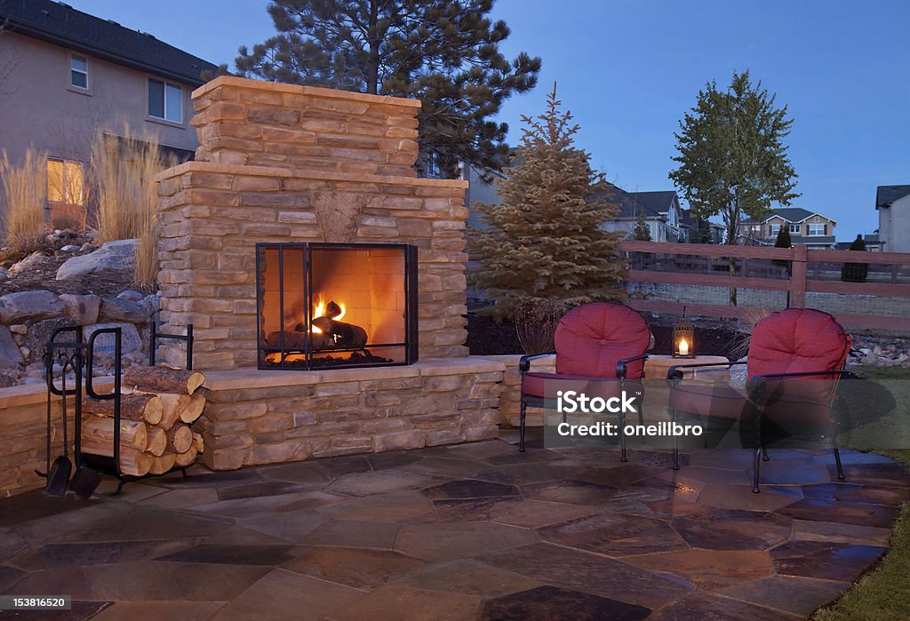 Outdoor flagstone platform with fireplace, chairs A backyard patio features a fireplace and casual furnishings.  The outdoor fireplace burns with a bright orange glow and is covered by a metal grate.  There is extra wood stored to the left of the stone fireplace in a black unit.  There are two metal chairs with red cushions placed on the flagstone patio. Fireplace Stock Photo