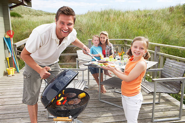 Family enjoying a porch barbecue on vacation There is a family with two children, one boy and one girl, on a wooden deck.  The dad is lifting up the lid of a grill that has burgers and vegetables cooking on it.  They are all smiling. family bbq beach stock pictures, royalty-free photos & images