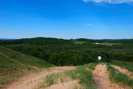 A woman takes in the view from atop the Sleeping Bear Dunes of Michigan. In the distance is sandy trail leading to the beach and a historic farm surrounded by green woods.