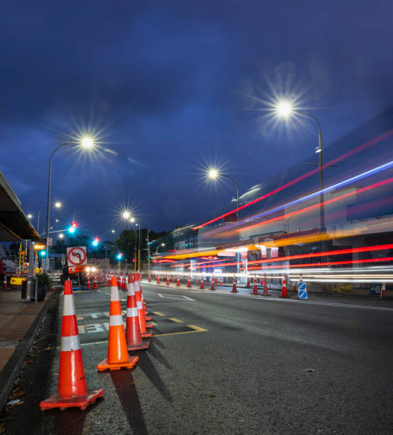 Orange traffic cones lining up the road. Bus light trails approaching the intersection. Roadworks in Auckland. Vertical format.