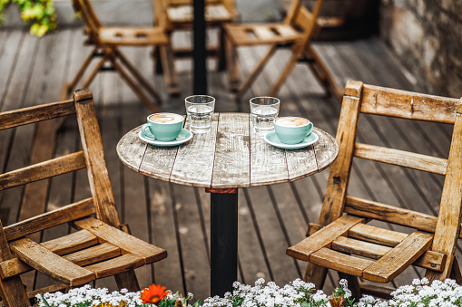 Two light blue cups of hot aroma coffee with latte art and glasses of water served on round wooden table at cafe. Beautiful summer terrace with chairs and flowers outdoors
