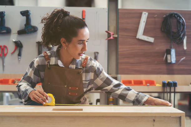 istock Young woman carpenter using measuring tape at carpentry workshop. Joiner female working with tape measure in wood factory 1538755964