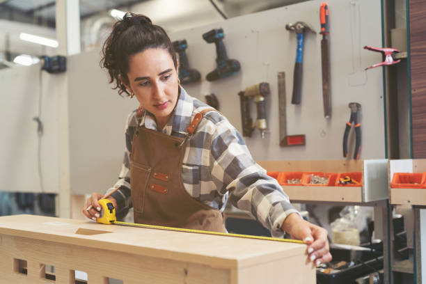 istock Young woman carpenter using measuring tape at carpentry workshop. Joiner female working with tape measure in wood factory 1538755972