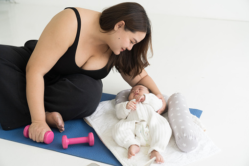 Happy smiling Asian young mother doing exercises on yoga mat and taking care her newborn at home