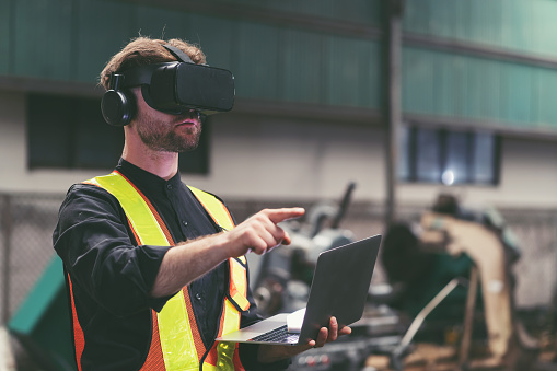Male factory mechanic using virtual reality headset. Male engineer working or using virtual reality headset for checking machinery in industry factory and wearing safety uniform and helmet