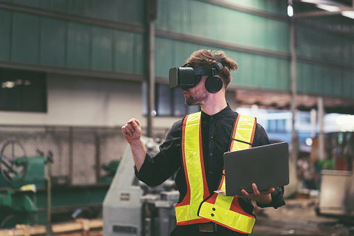 Male factory mechanic using virtual reality headset. Male engineer working or using virtual reality headset for checking machinery in industry factory and wearing safety uniform and helmet