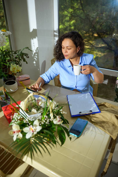 Multitasking woman florist event manager drinks coffee, working on tablet, checking online orders in her comfy office