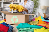 The concept of recycling clothes. A woman puts bright colored clothes in a box with a recycle sign.