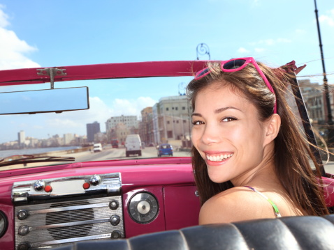 Car xwoman smiling happy on passenger seat in pink vintage convertible car. Young mixed race Asian / Caucasian female model during cuba vacation. Driving on Malecon road in Old Havana.