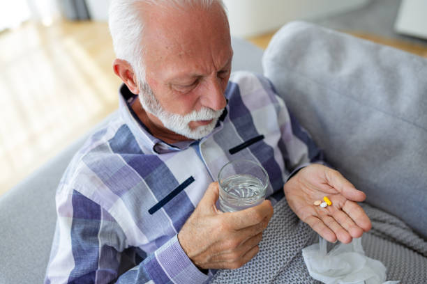 istock Senior man taking pills while sitting on couch at home, holding white jar with treatment, copy space. Grey-haired elderly man using supplements or vitamins. Healthy lifestyle in senior age concept 1538992219