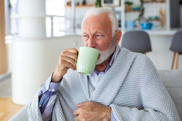 istock Senior man suffering from flu drinking tea while sitting wrapped in a blanket on the sofa at home. Sick Man with a cold lying on the sofa holding a mug of hot tea. 1538992226