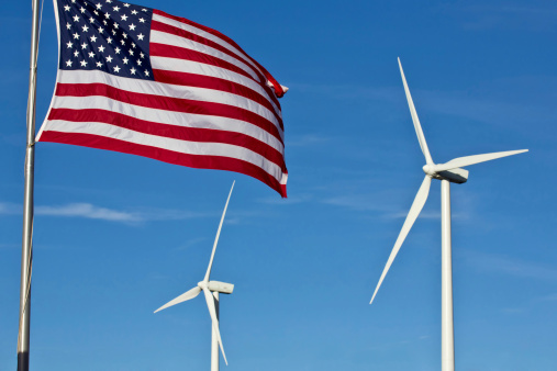 An american flag flies in the foreground, with two wind power generator windmills turn in the background