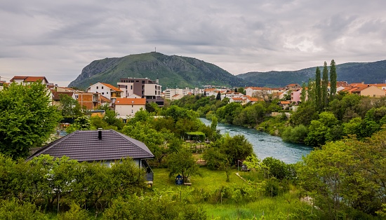 Mostar, Bosnia and Herzegovina – May 10, 2023: An aerial view of a quaint, small-town nestled between mountain ranges and a winding river, with cloudy skies in the background