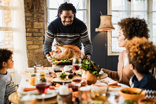 Happy African American father bringing Thanksgiving turkey to the table for his family's' lunch.