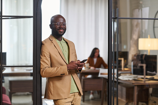 Happy young African American male designer in formalwear looking at camera while standing by open door of office and using smartphone