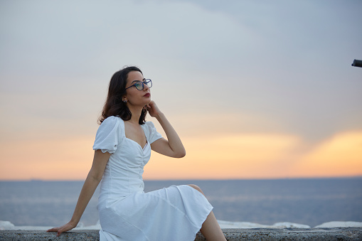 Attractive young brunette lady  with short hair and eyeglasses sitting over rocks near seaside during sunset