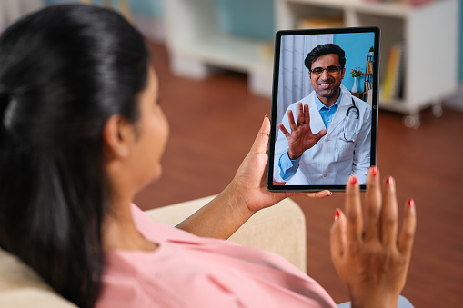 Shoulder shot of indian pregnant woman consulting doctor on video call at home - concept telemedicine, Virtual healthcare and remote consultation