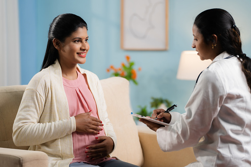 Indian doctor or nurse counseling pregnant woman at home by writing report at home - concept of home health checkup, maternity assistance and Medicare support