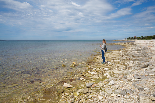 Senior woman standing on the edge of Adriatic sea and looking far out at horizon. She is in stones and sandy beach, sea water in front of her and beautiful blue sky with white clouds. It is early spring near Fazana, Istrian coast in Croatia.