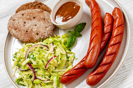 close-up of juicy cooked sausages with classic coleslaw salad, bbq sauce and rye bread on plate on white wood table