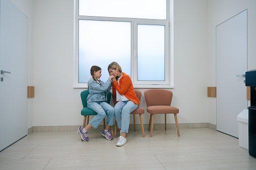 Girl with her mother are sitting on armchairs in a bright hospital corridor, a woman kisses the girls hands