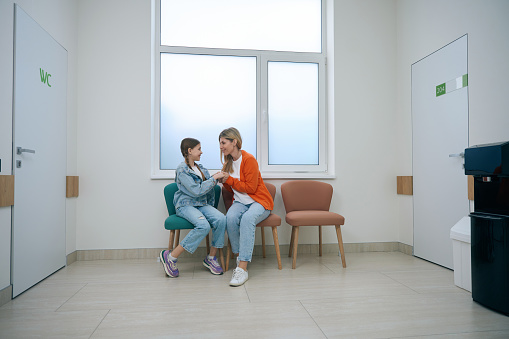 Cheerful girl with her mother are sitting on armchairs in bright hospital corridor, a woman is holding the girls hands