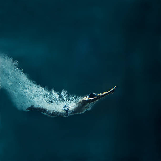 Swimmer diving after the jump, underwater view The male swimmer has straight body. The image is made just after entering the water. His body is covered by air bubbles. Hands are joined in front of the body. He wears black cap and black swimwear. The swimmer has muscular built. He is leaving behind a lot of air bubbles. The background is dark blue. There are not visible parts of swimming pool. There is a lot of free space around the swimmer. This is a square format photography. diving into water stock pictures, royalty-free photos & images