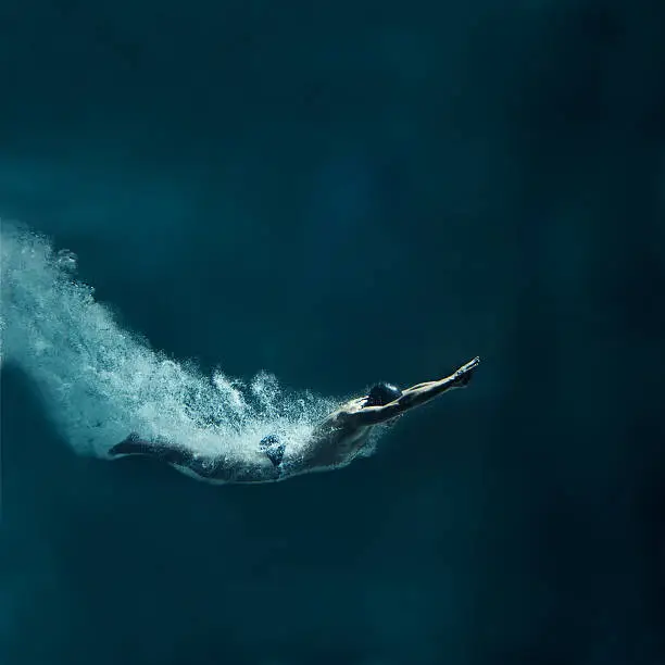 The male swimmer has straight body. The image is made just after entering the water. His body is covered by air bubbles. Hands are joined in front of the body. He wears black cap and black swimwear. The swimmer has muscular built. He is leaving behind a lot of air bubbles. The background is dark blue. There are not visible parts of swimming pool. There is a lot of free space around the swimmer. This is a square format photography.