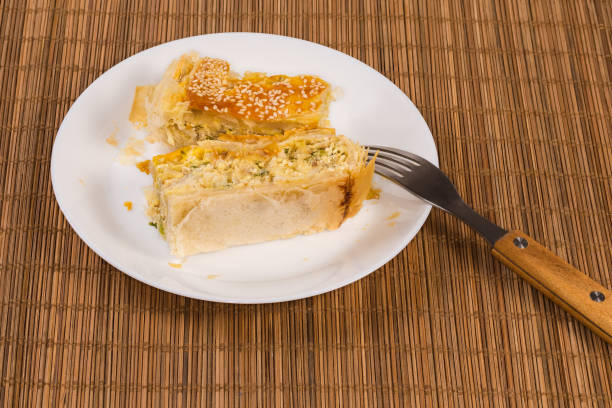 Pieces of the pie made with puff pastry with savory fish filling, sprinkled with sesame on the white dish with fork on the bamboo place mat