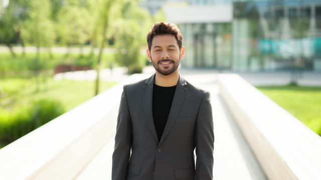Cheerful young corporate looks at camera and smiles. Happy handsome guy standing outside a modern building smiling and looking straight at camera
