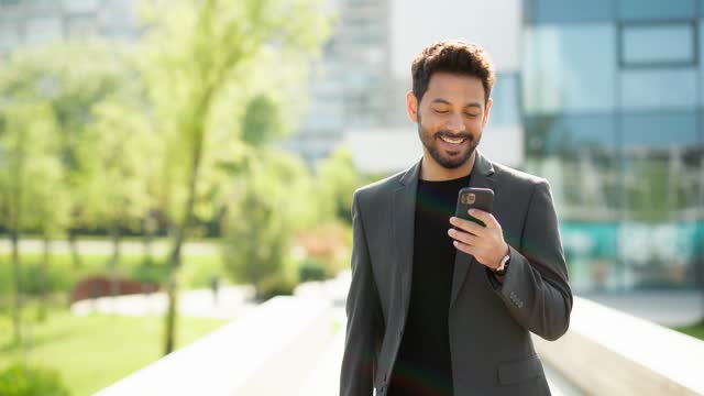 Handsome young entrepreneur using a smartphone and smiling. Attractive cheerful businessman walking outside a modern building and scrolling internet, social media, news using his phone
