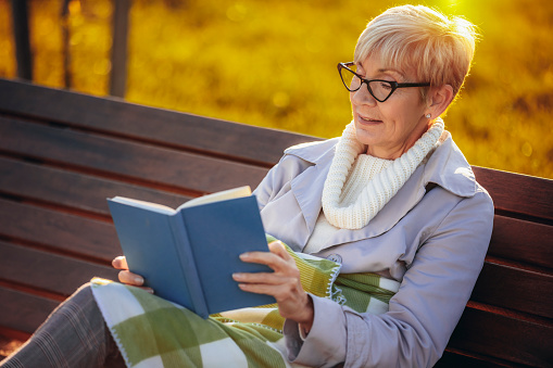 An older woman reading a book. She is sitting on the bench in the colorful park in the autumn