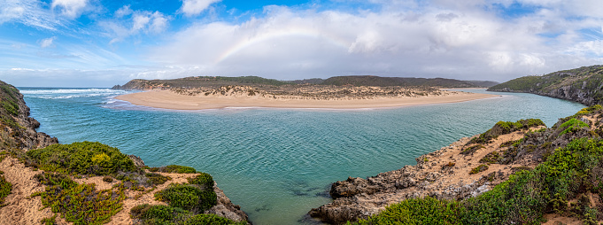 Ultra wide panorama of a rainbow in front of the clouds at Praia da Amoreira, one of the surfers' favorite beach with beautiful wide sandy beach, long waves, river and cliffs, Algarve, Portugal