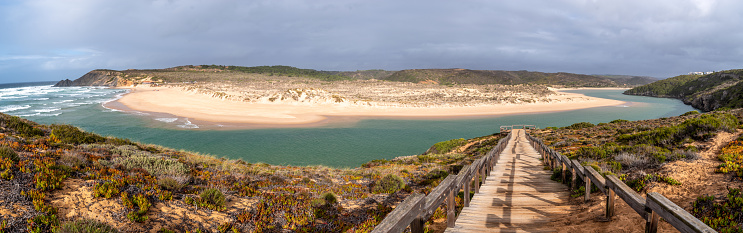 Ultra wide angle panorama of Wooden walkway to the beach Praia da Amoreira at river Ribeira in Aljezur, snaking through the golden sand of Praia da Amoreira beach with crystal clear calm water, sand dunes and clouds in the sky on a sunny day in October.