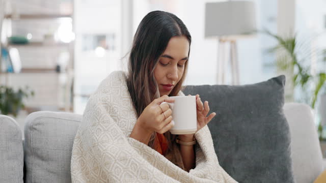 Woman, drink and tea in home with blanket for comfortable, relaxing and carefree weekend on living room sofa. Female person drinking cup of hot beverage, coffee and mug to stay warm, cosy and rest